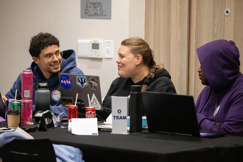 Three hackathon participants sit at a table with laptops open, collaborating and discussing solutions.